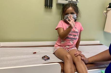 Three-year-old girl sits on exam table in clinic waving to camera about to get vaccination
